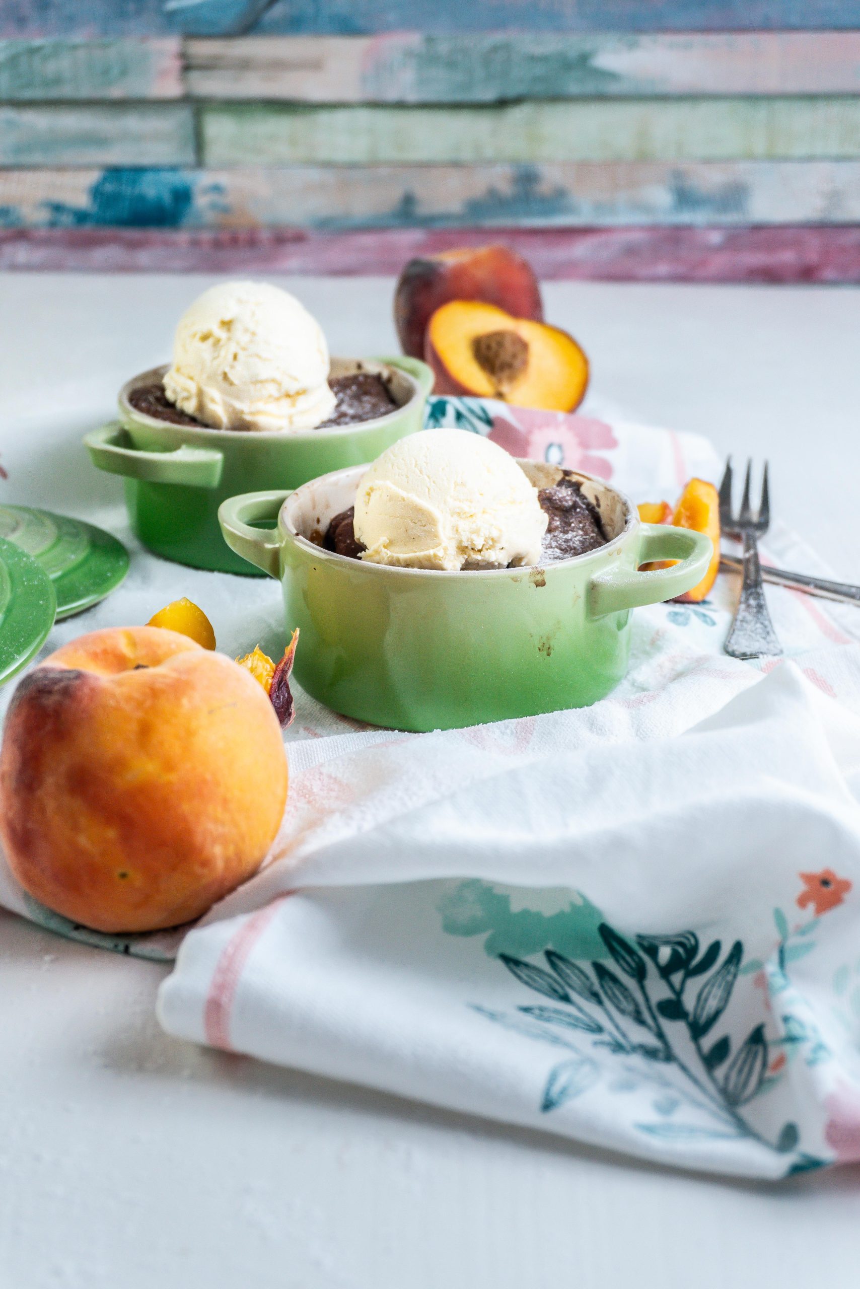 Mini Peach Cobblers with ice cream on top in small ramekins with peaches strewn around and two forks lying on the side; photographed from a side-angle