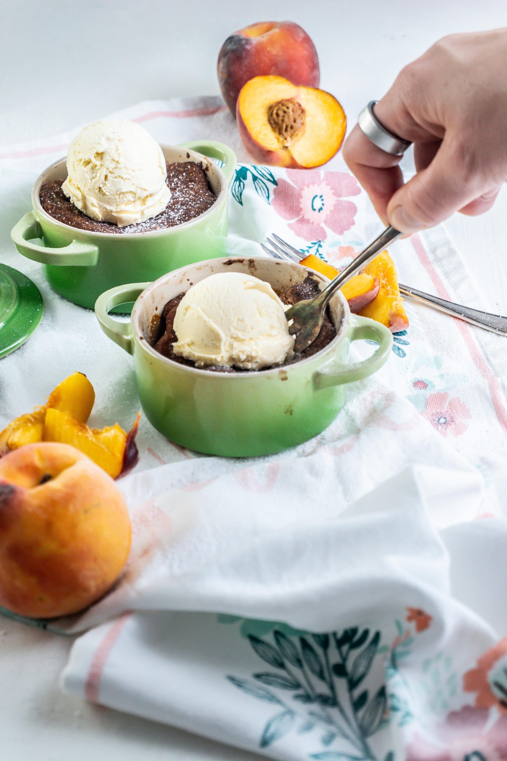 Mini Peach Cobblers with ice cream on top in small ramekins with peaches strewn around and a fork digging into one of the cobblers