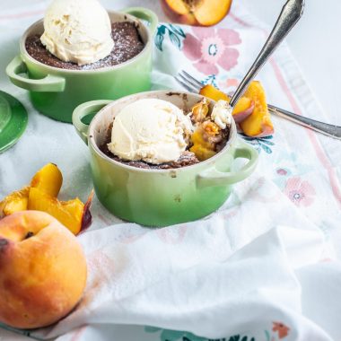 Mini Peach Cobblers with ice cream on top in small ramekins with peaches strewn around, and a fork being inside the ramekin dish