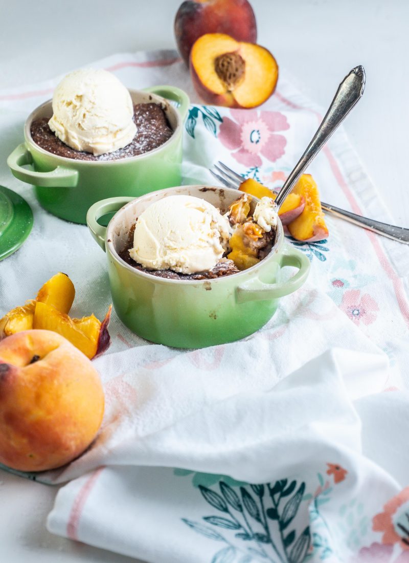Mini Peach Cobblers with ice cream on top in small ramekins with peaches strewn around, and a fork being inside the ramekin dish