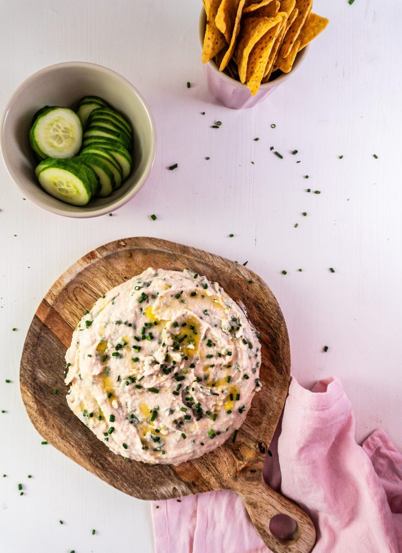 Quick and Easy Butterbean Dip on a wooden plate with cucumbers and nachos visible in the background and garnished with chives