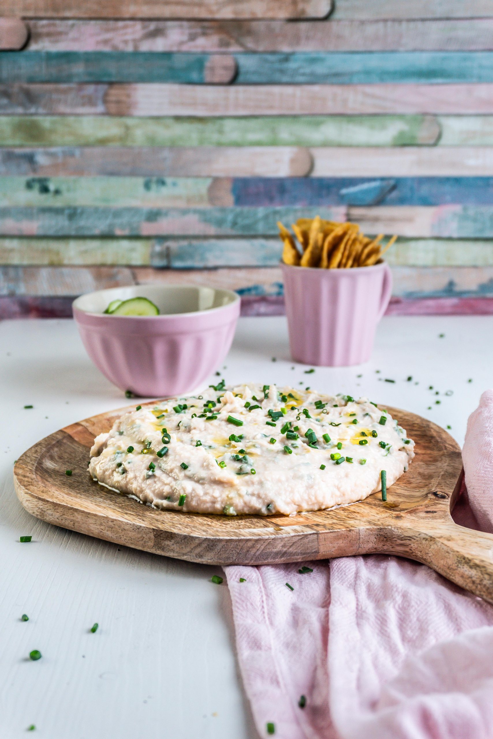Quick and Easy Butterbean Dip on a wooden plate with cucumbers and nachos visible in the background and garnished with chives; photographed from a side angle